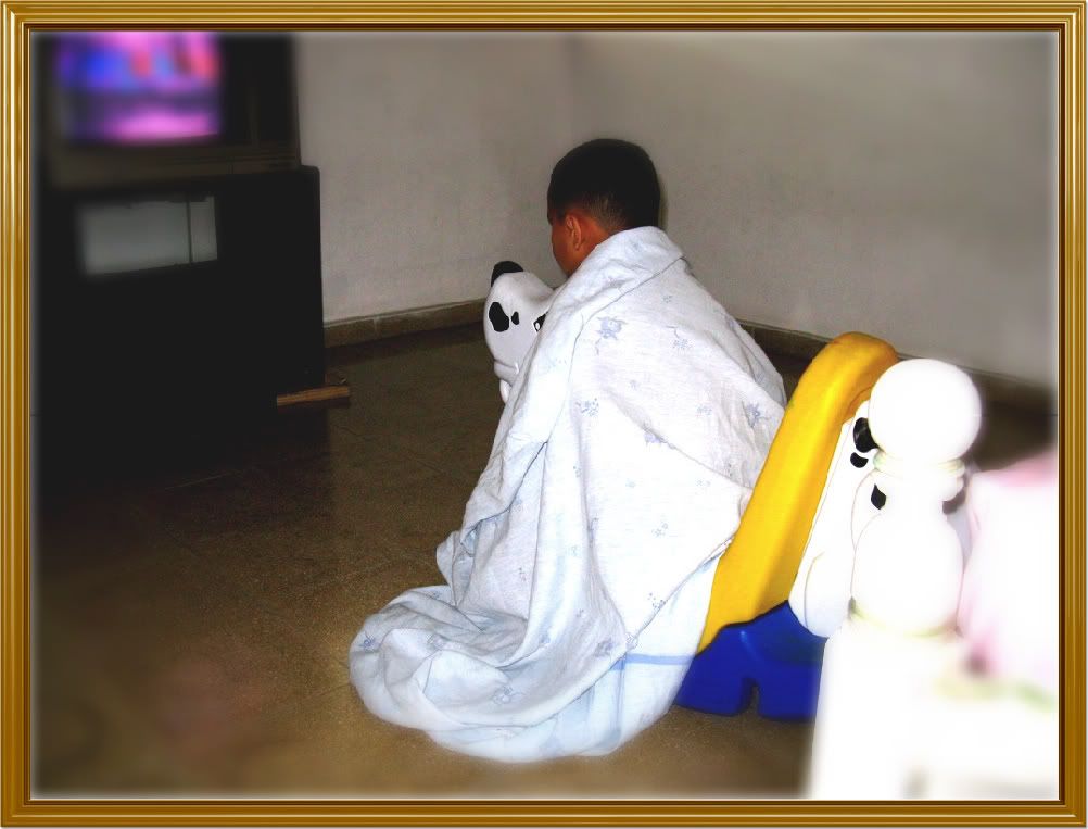 Photograph of Abdiel rocking on a plastic dog, wrapped up in a sheet and with his eyes glued to the screen of the TV set.