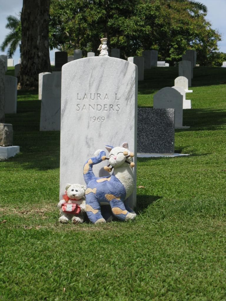 Photograph of the tombstone of Laura L. Sanders with two toys poised above the lawn. (Credit: Omar Upegui R.)